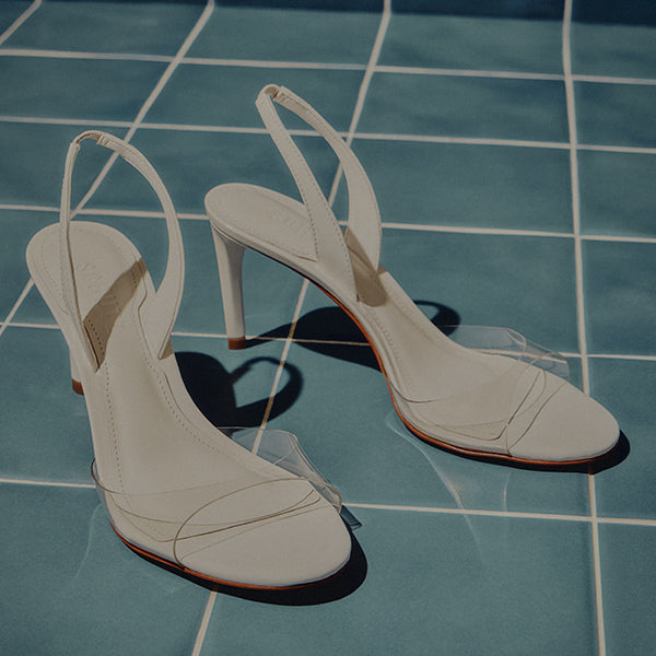 Elegant white high-heeled sandals with clear straps are placed on blue tiled flooring, casting soft shadows, conveying a chic and minimalist style.
