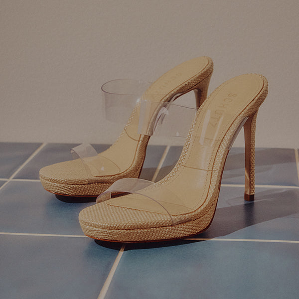 Beige high-heeled sandals with clear plastic straps on a blue tiled surface. The lighting creates a soft, elegant tone, highlighting the shoe texture.