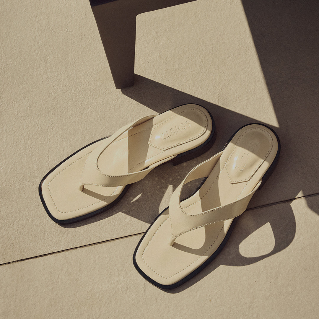 Beige leather sandals with black soles rest on sunlit tiles, casting soft shadows. The scene conveys a warm, relaxed, summery feel.