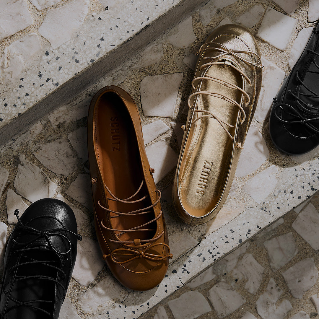 Three ballet flats on tiled steps; one black, one brown, and one metallic gold. Each shoe features delicate bow laces, creating a stylish and elegant feel.