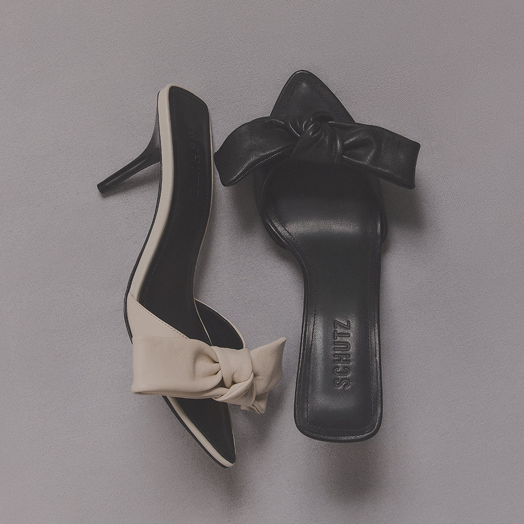 Chic high-heeled mules on a gray surface, with one shoe in cream and the other in black, both featuring elegant bows, signifying modern elegance.