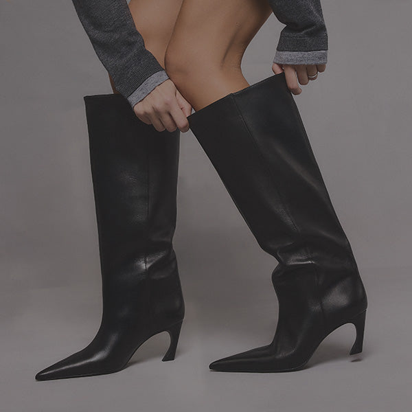 A person wearing tall, sleek black leather boots with pointed toes and low heels adjusts them. They are dressed in a gray outfit, conveying a chic, modern look.