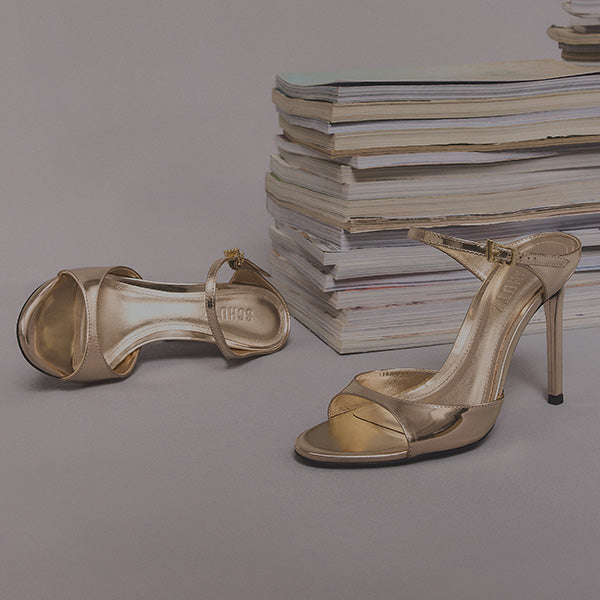 A pair of elegant gold high-heeled sandals placed in front of a stack of magazines. The setting conveys a sophisticated and stylish tone.