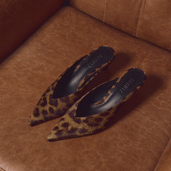 Leopard print pointed-toe mules with black interiors sit stylishly on a rich brown leather surface, conveying a chic and luxurious feel.