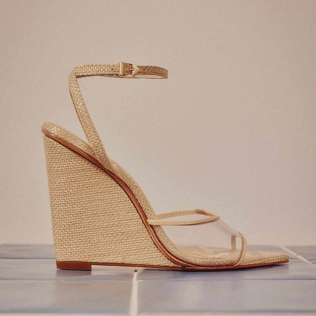 A beige woven wedge sandal with a clear strap and ankle buckle, placed on blue tiles. The style is elegant and summery, exuding a chic tone.