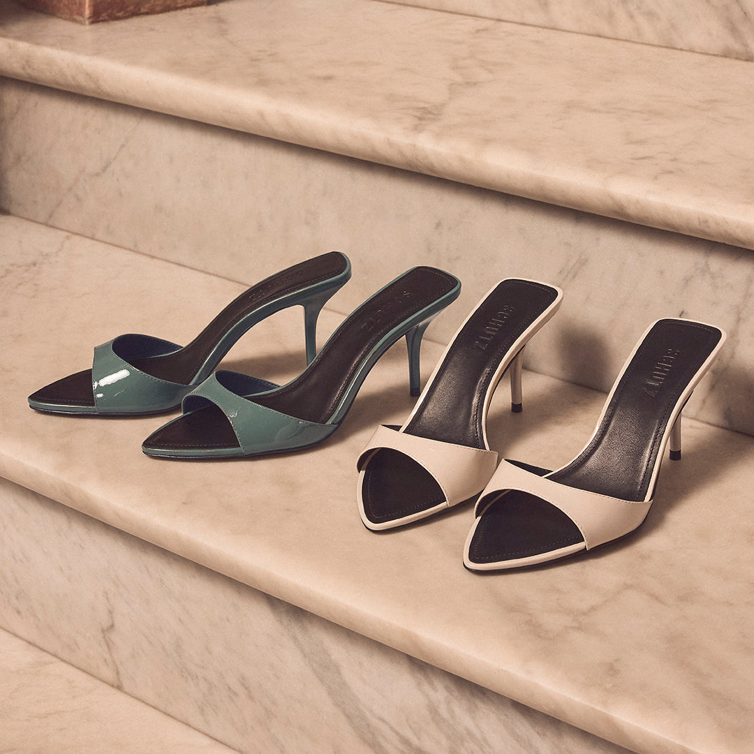Two pairs of sleek high-heeled mules on marble stairs; one teal and one beige, exuding elegance and sophistication in a soft, warm light.