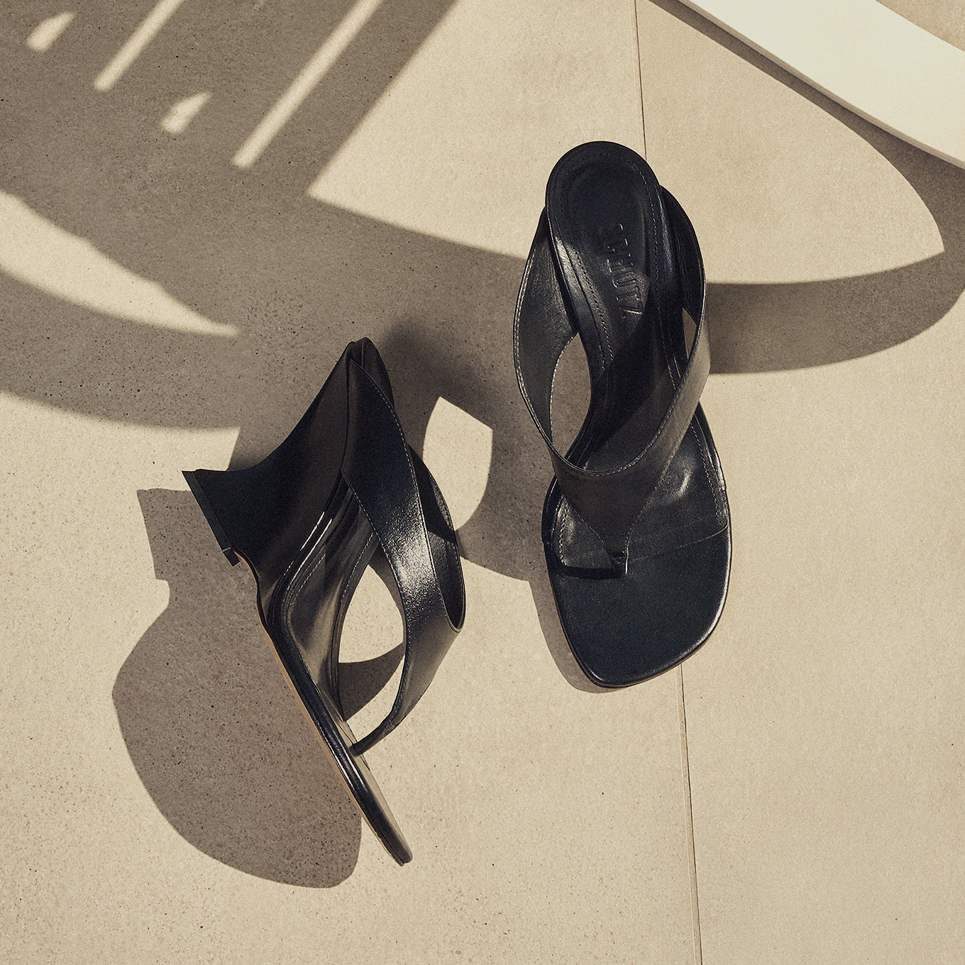 Black high-heeled sandals with wide straps placed on a sunlit tiled surface. The geometric shadows cast create an elegant, sophisticated tone.
