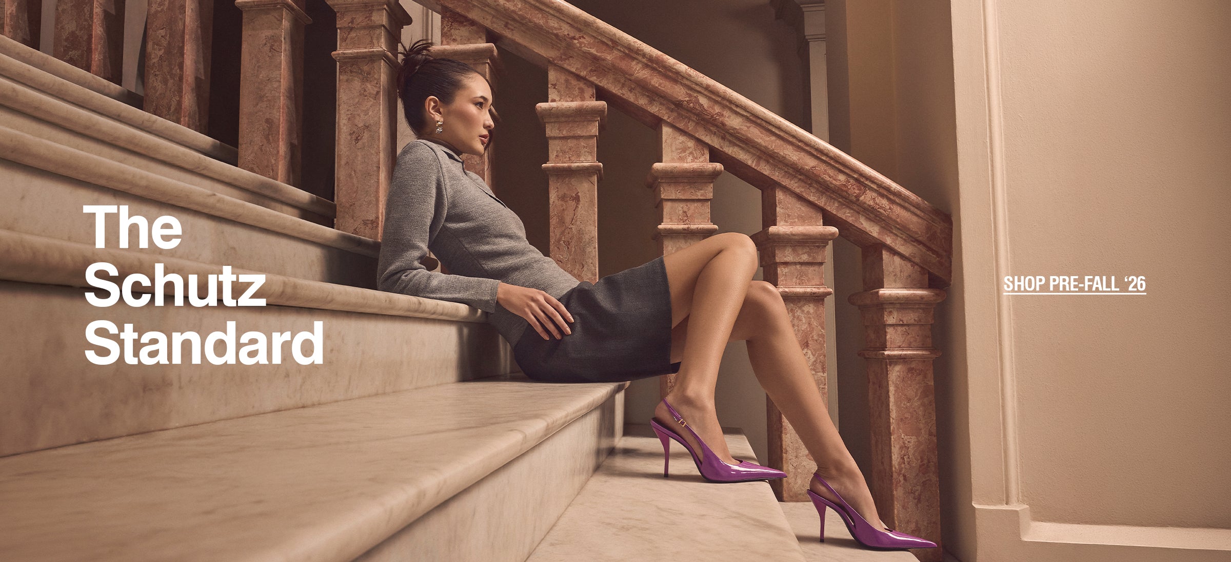 A woman in a gray top and dark skirt reclines on marble stairs, wearing purple heels. Text reads "The Schutz Standard" and "Shop Pre-Fall '26."