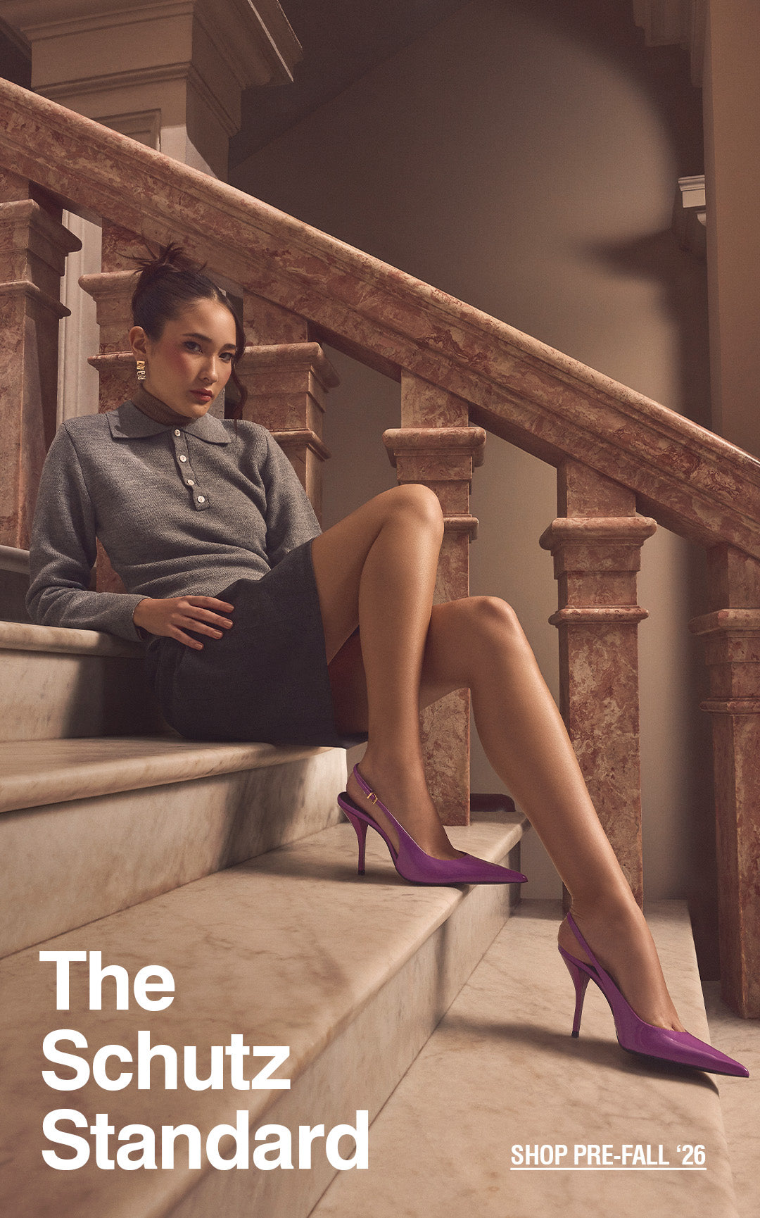 A woman in a gray top and dark skirt reclines on marble stairs, wearing purple heels. Text reads "The Schutz Standard" and "Shop Pre-Fall '26."
