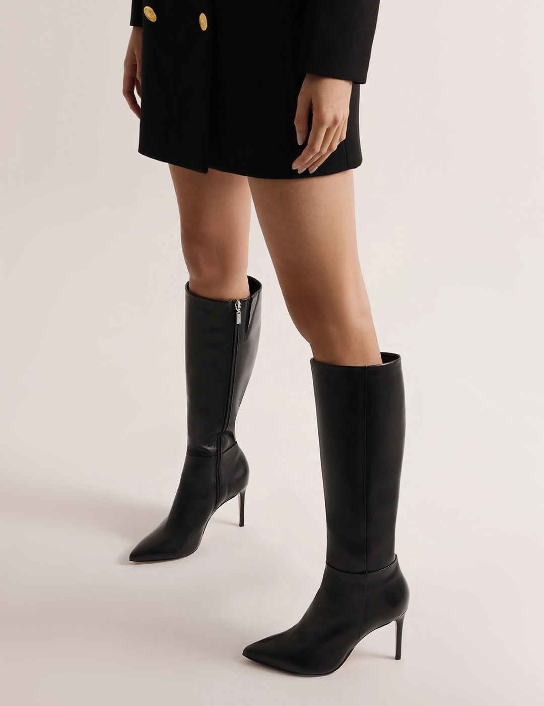 A person wearing black knee-high pointed-toe heeled boots with side zippers, paired with a black blazer, shown against a plain light beige studio background.