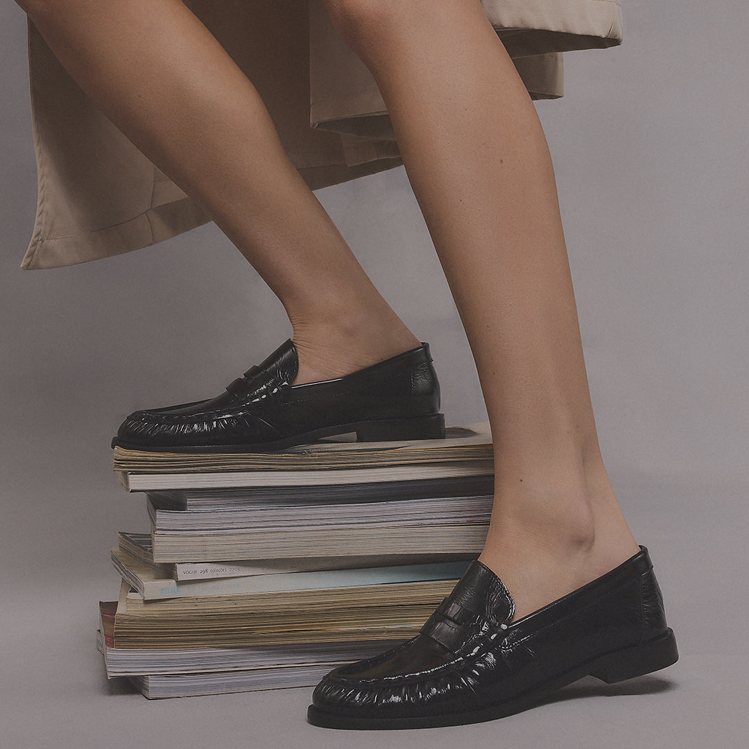Legs in a light skirt, wearing black patent leather loafers, with one foot on a stack of magazines and the other on the floor.