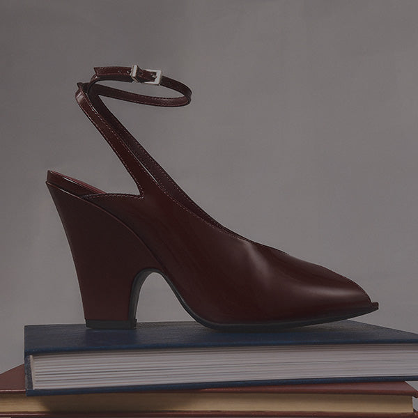 Dark red patent leather pointed-toe slingback heel with an ankle strap, resting on a stack of books.