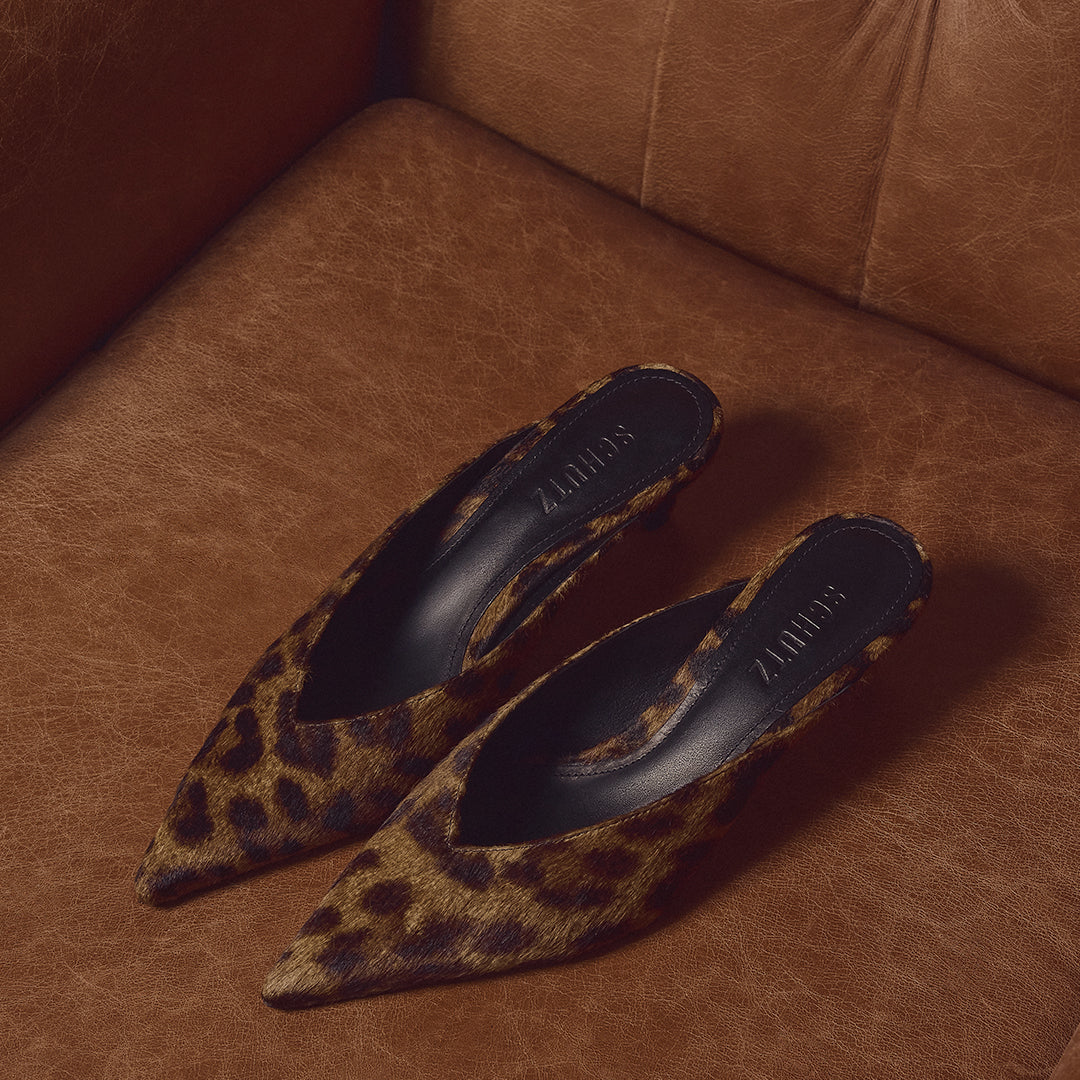 A pair of pointed-toe mule heels in a brown and black leopard print, displayed on a brown leather sofa. The brand name 'SCHUTZ' is visible on the inner sole.