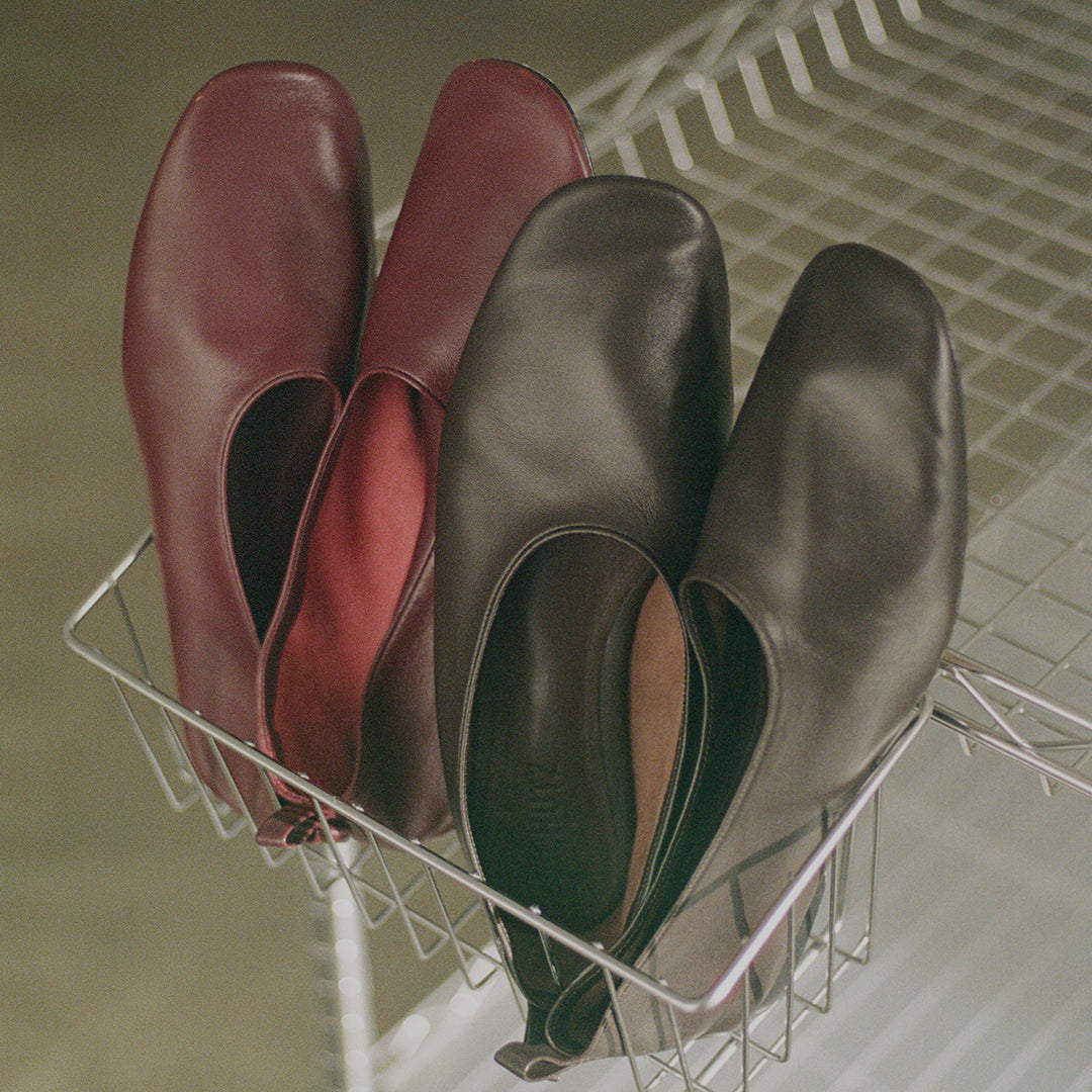 Four slip-on flats in burgundy and charcoal gray leather, arranged in a wire basket.
