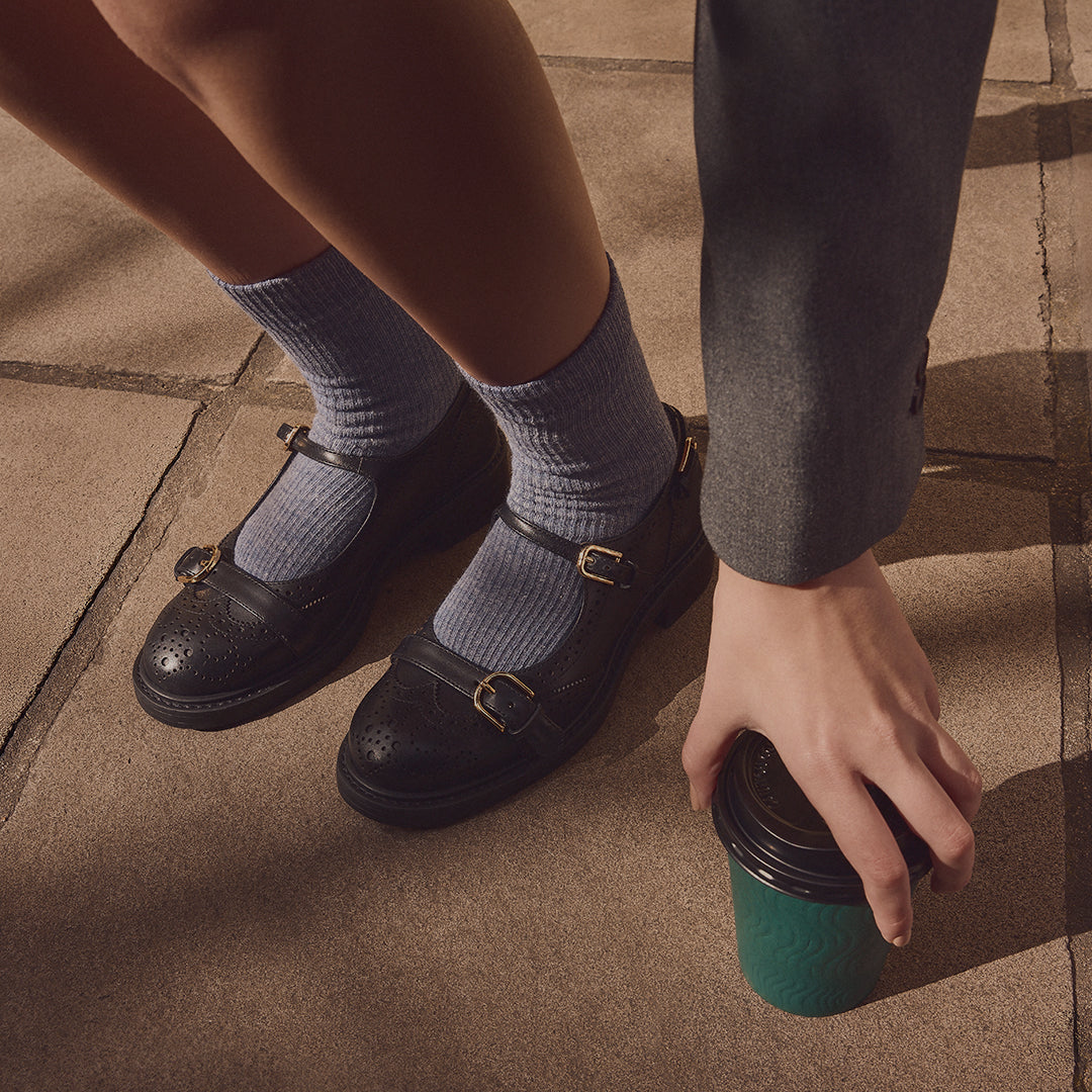 A person wearing black Mary Jane style loafers with gold-tone buckles, light blue ribbed socks, and a dark gray blazer, holding a green paper coffee cup on a stone pavement.