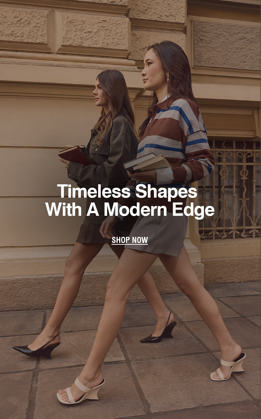 Two women walking outdoors in front of a beige stone building. The woman in front wears a brown and blue striped knit sweater, a gray skirt, and beige heeled sandals, while the woman behind wears a dark olive jacket and shorts with black heels.