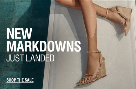 A model's legs wearing beige raffia strappy wedge sandals are shown against a split background of dark teal tiles and a light gray floor, with promotional text reading 'NEW MARKDOWNS JUST LANDED' and 'SHOP THE SALE'.