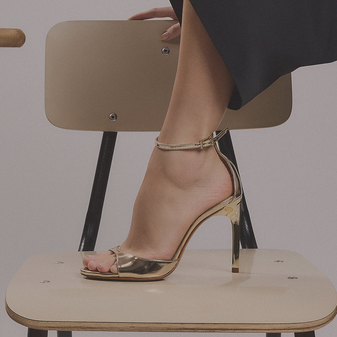 A close-up of a person's foot wearing a metallic gold high-heeled sandal with an ankle strap, seated on a minimalist beige chair.