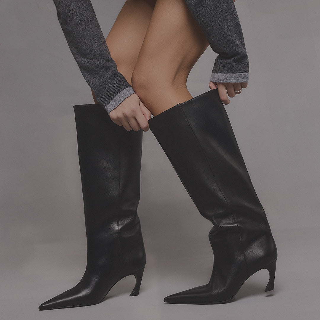 A person wearing black pointed-toe knee-high heeled boots, shown from the waist down against a plain gray studio background.