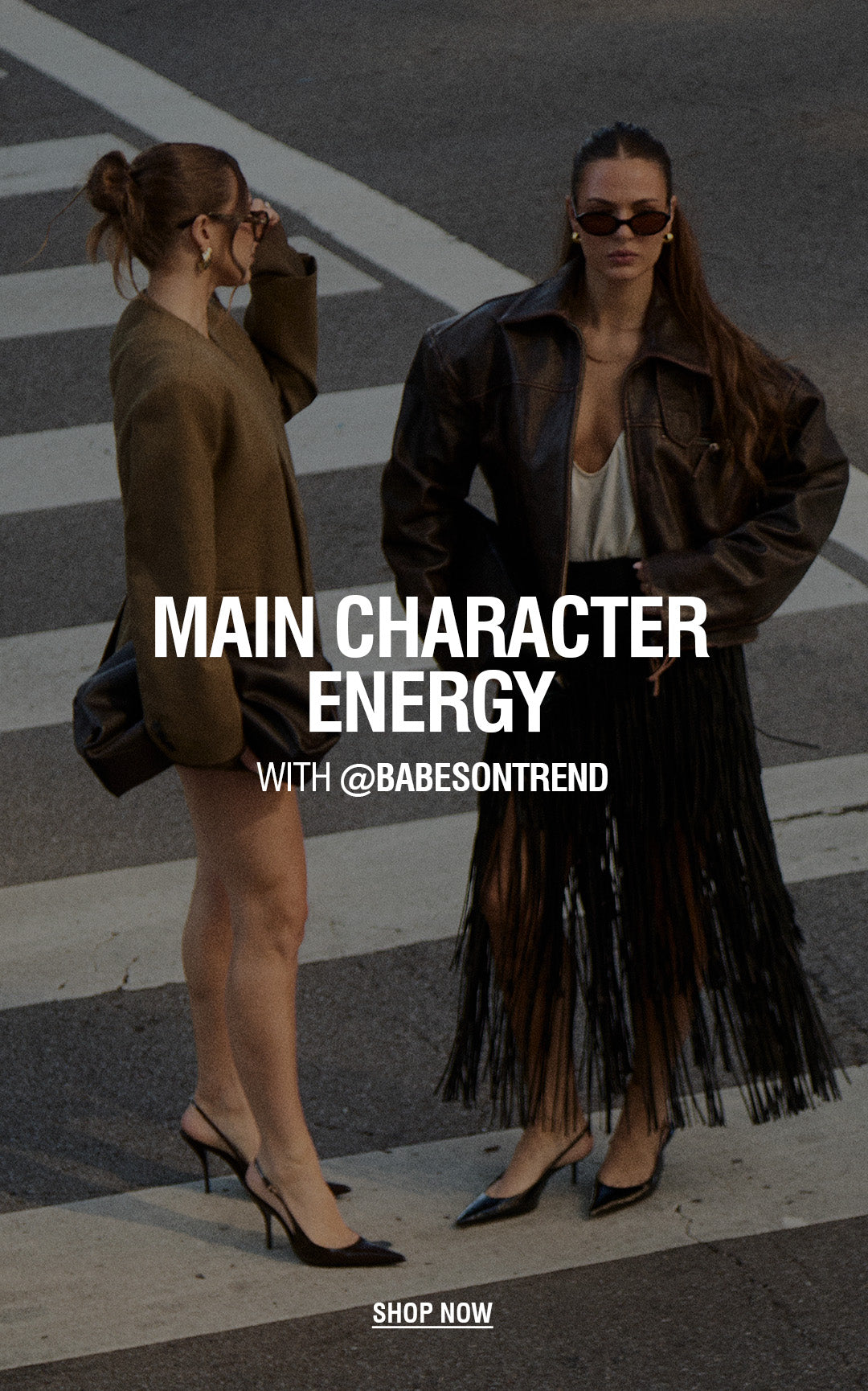 Two women standing on a street crosswalk wearing streetwear fashion. One wears an oversized brown blazer and black heels, while the other wears a black leather jacket with a long black fringe skirt over a white top and black heels. Text Reads: MAIN CHARACTER ENERGY with @babesontrend. Shop Now.