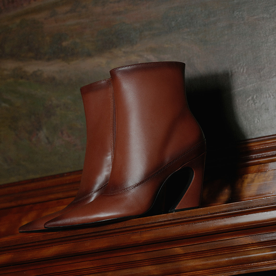 A pair of brown leather ankle boots with pointed toes and sculptural block heels, displayed on a dark wooden mantelpiece against a textured wall.