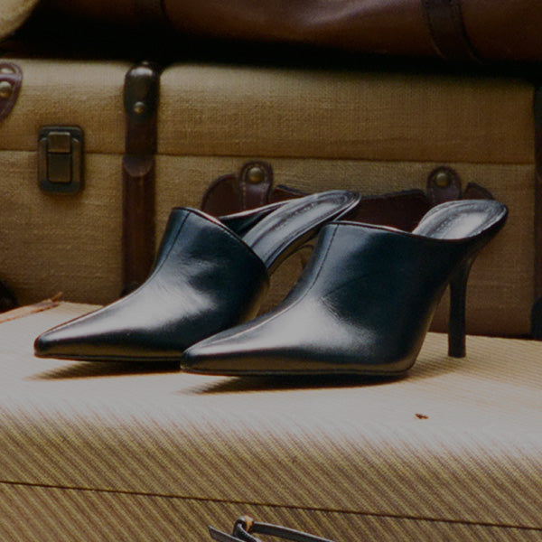 A pair of black leather pointed-toe mule heels with mid-height stiletto heels, displayed on a textured beige suitcase against a vintage brown suitcase background.