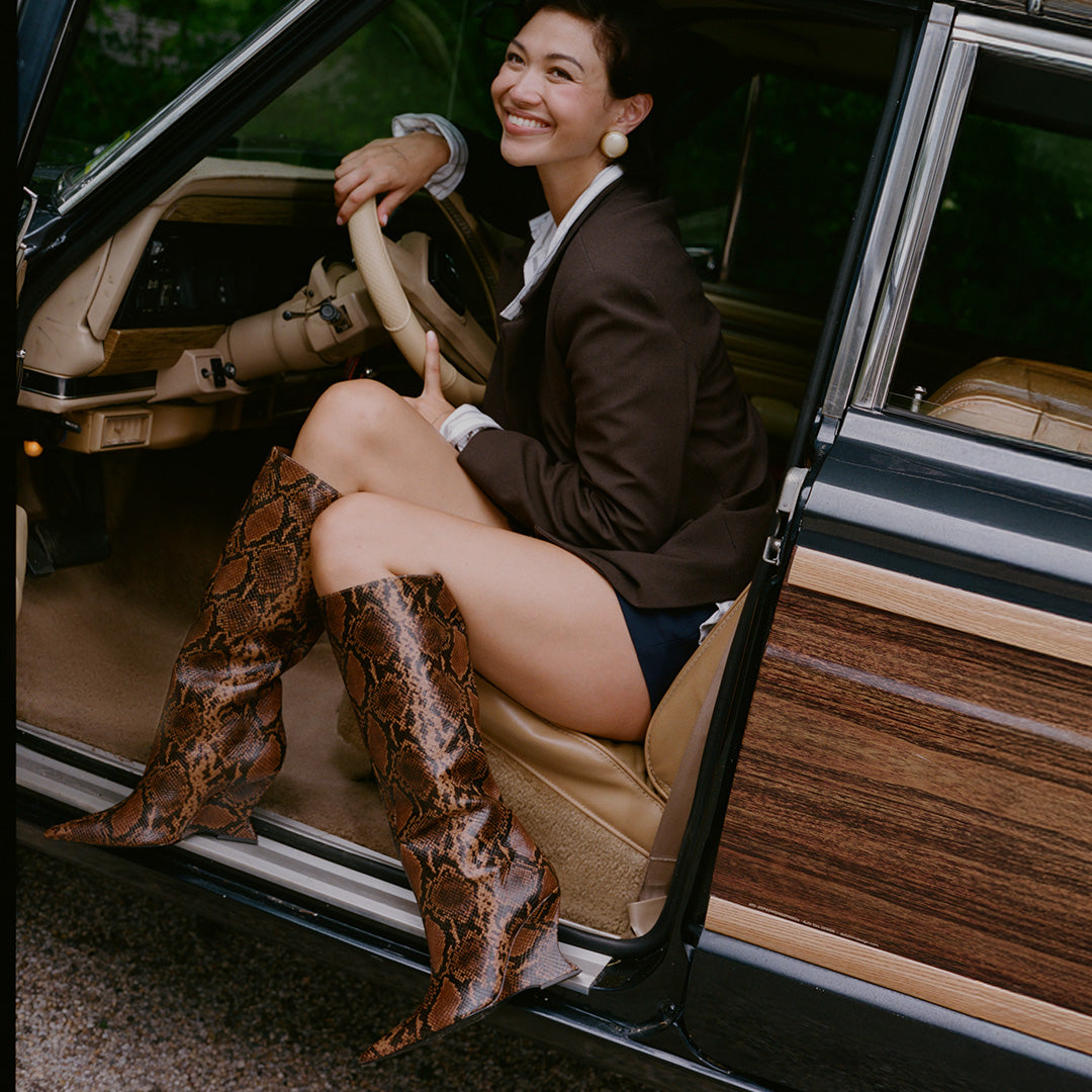 A woman sitting in the open door of a vintage wood-paneled car, wearing brown snakeskin-patterned knee-high boots, a dark brown blazer, a white collared shirt, and dark shorts.