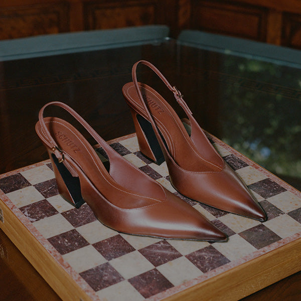 A pair of pointed-toe slingback pumps in smooth cognac brown leather, displayed on a checkered chessboard with a dark wooden background.