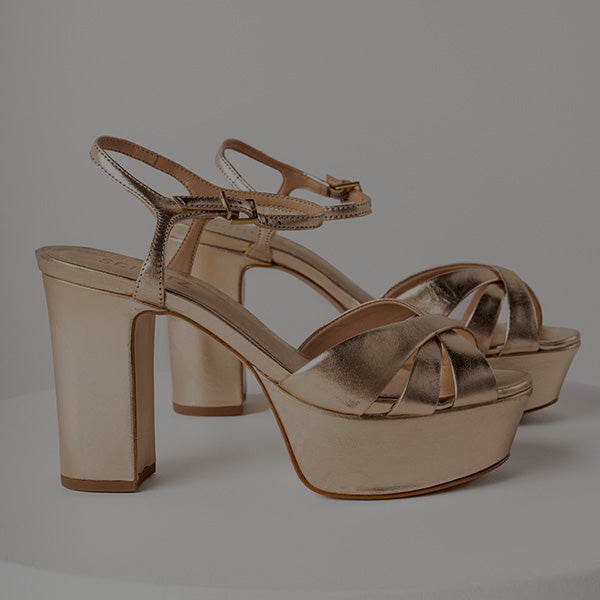 A pair of metallic gold platform high-heeled sandals with crisscross front straps and ankle straps, shown on a neutral gray background.