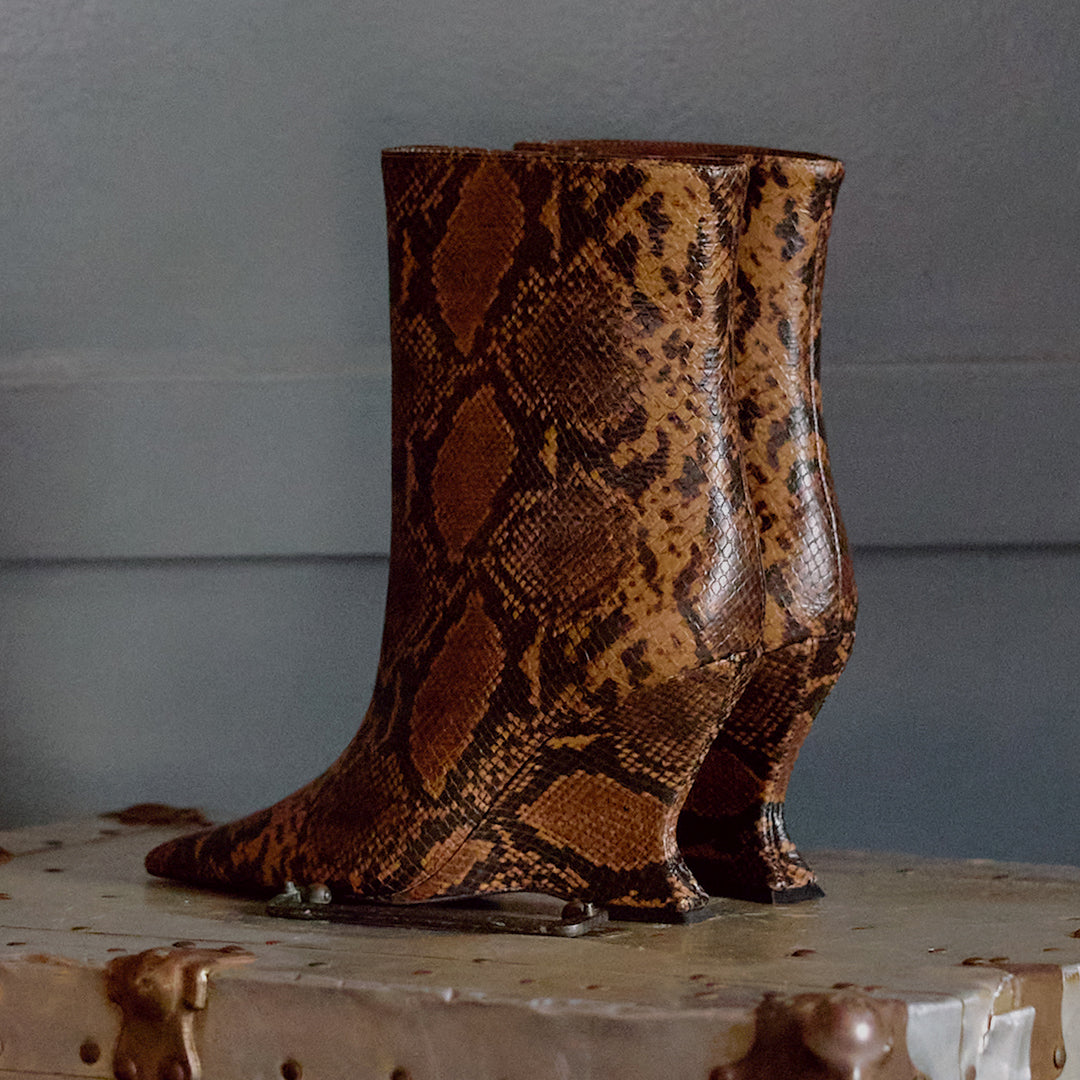 A pair of mid-calf boots with a brown and tan snakeskin pattern, displayed on a rustic metal surface against a gray paneled wall.