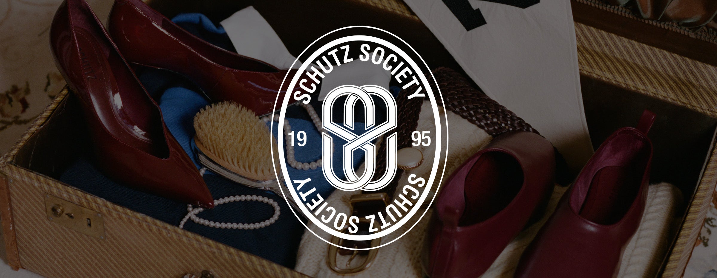 A vintage-style open suitcase filled with burgundy high-heeled shoes, a blue scarf, a wooden brush, and a pearl necklace, with a large circular Schutz Society logo overlay in the center.