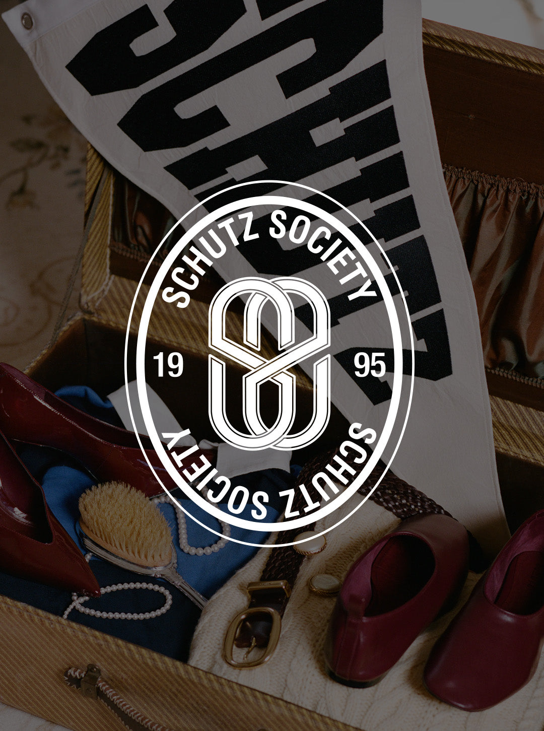 Schutz Society logo overlay over a styled flat-lay of a vintage suitcase containing burgundy shoes, a blue scarf, and a white bag with black lettering.