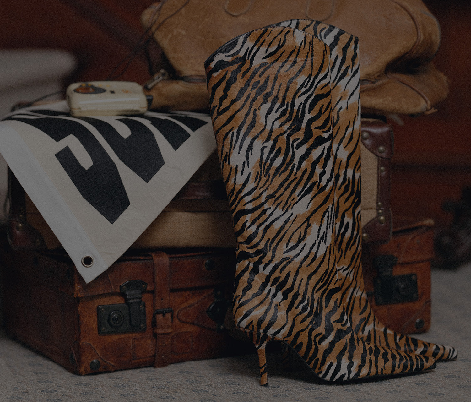 A tall pointed-toe heeled boot with a bold tiger-stripe pattern in brown, black, and white, styled on top of vintage brown leather luggage.
