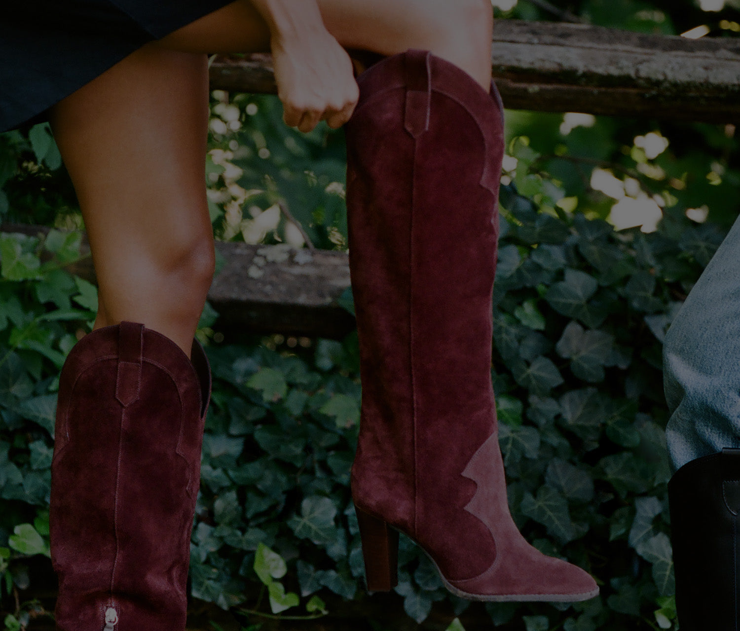 A person sitting outdoors on a wooden fence wearing tall burgundy suede cowboy boots with mid-calf counterparts visible. The background is filled with green ivy.