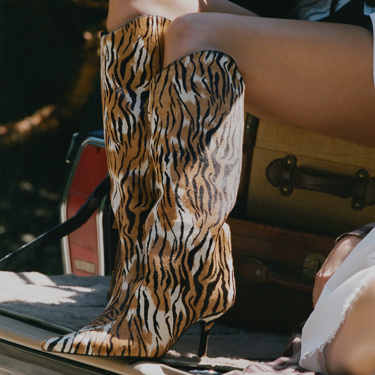 A close-up of a person wearing a tall pointed-toe heeled boot with a bold tiger-stripe pattern in tan, black, and cream, seated in a rustic setting with a suitcase in the background.