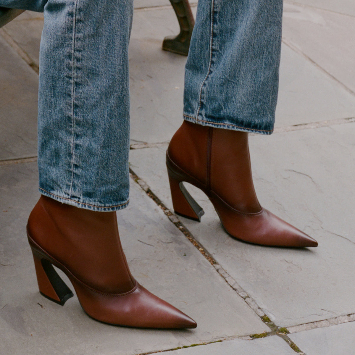 A person wearing brown leather pointed-toe ankle boots with block heels, paired with blue jeans, standing on a stone paved surface.
