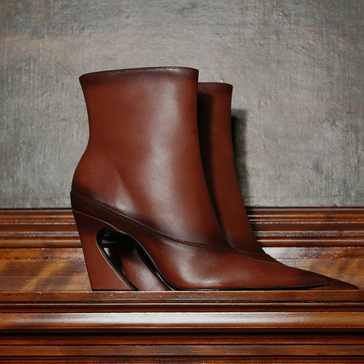 A pair of dark brown leather ankle boots with pointed toes and sculptural wedge heels, displayed on a polished wooden surface against a textured gray wall.