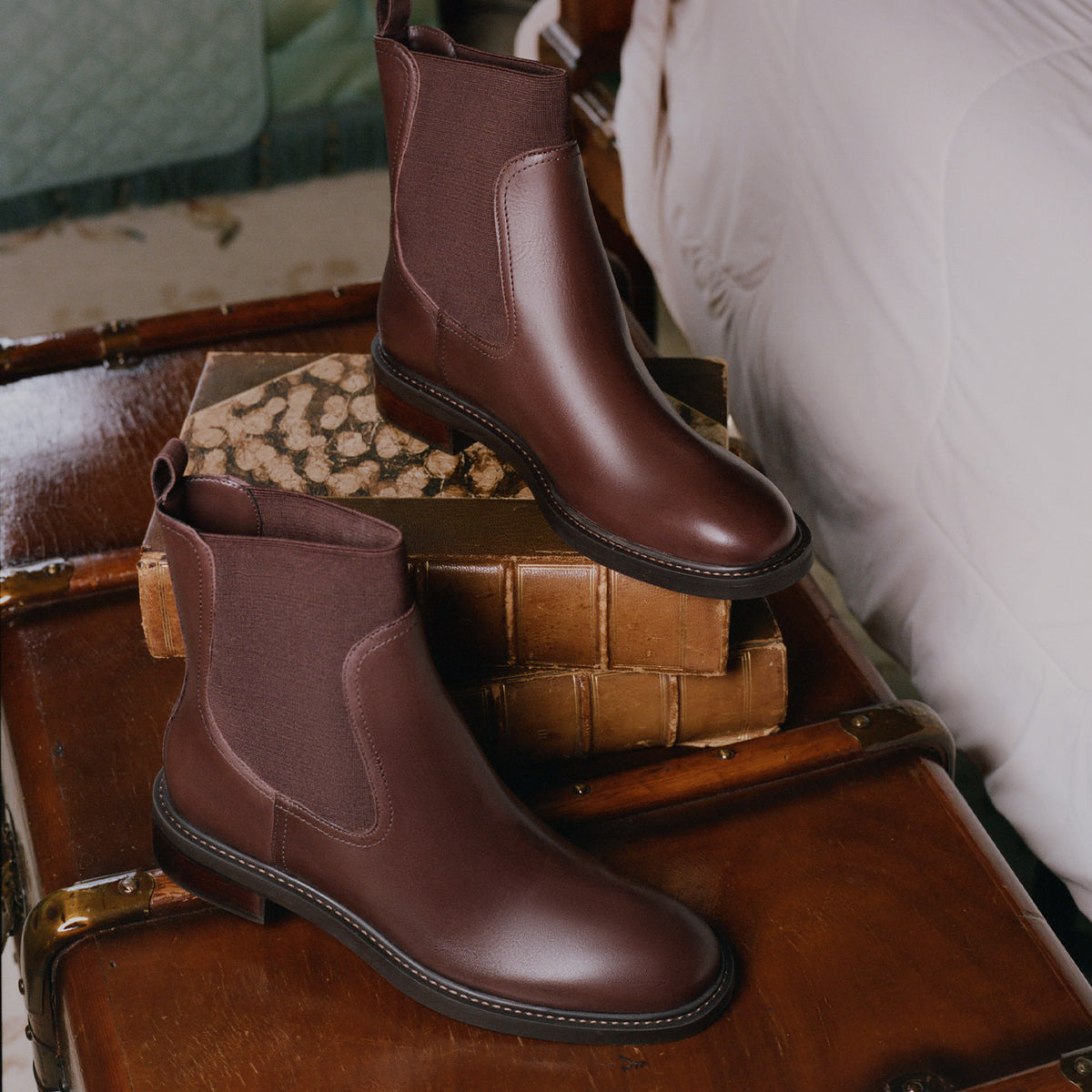A pair of dark brown leather Chelsea boots styled on vintage books and a wooden trunk next to a white upholstered chair.