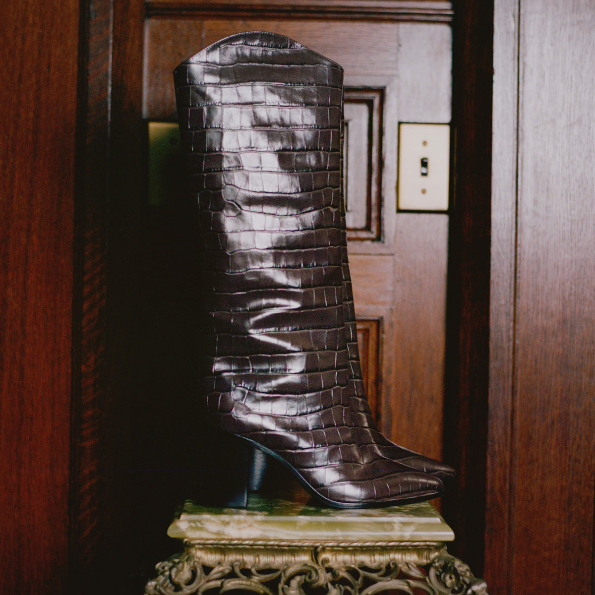 A tall, dark brown crocodile-embossed leather boot with a pointed toe and slim heel, displayed on an ornate stone pedestal in front of a dark wood-paneled wall.