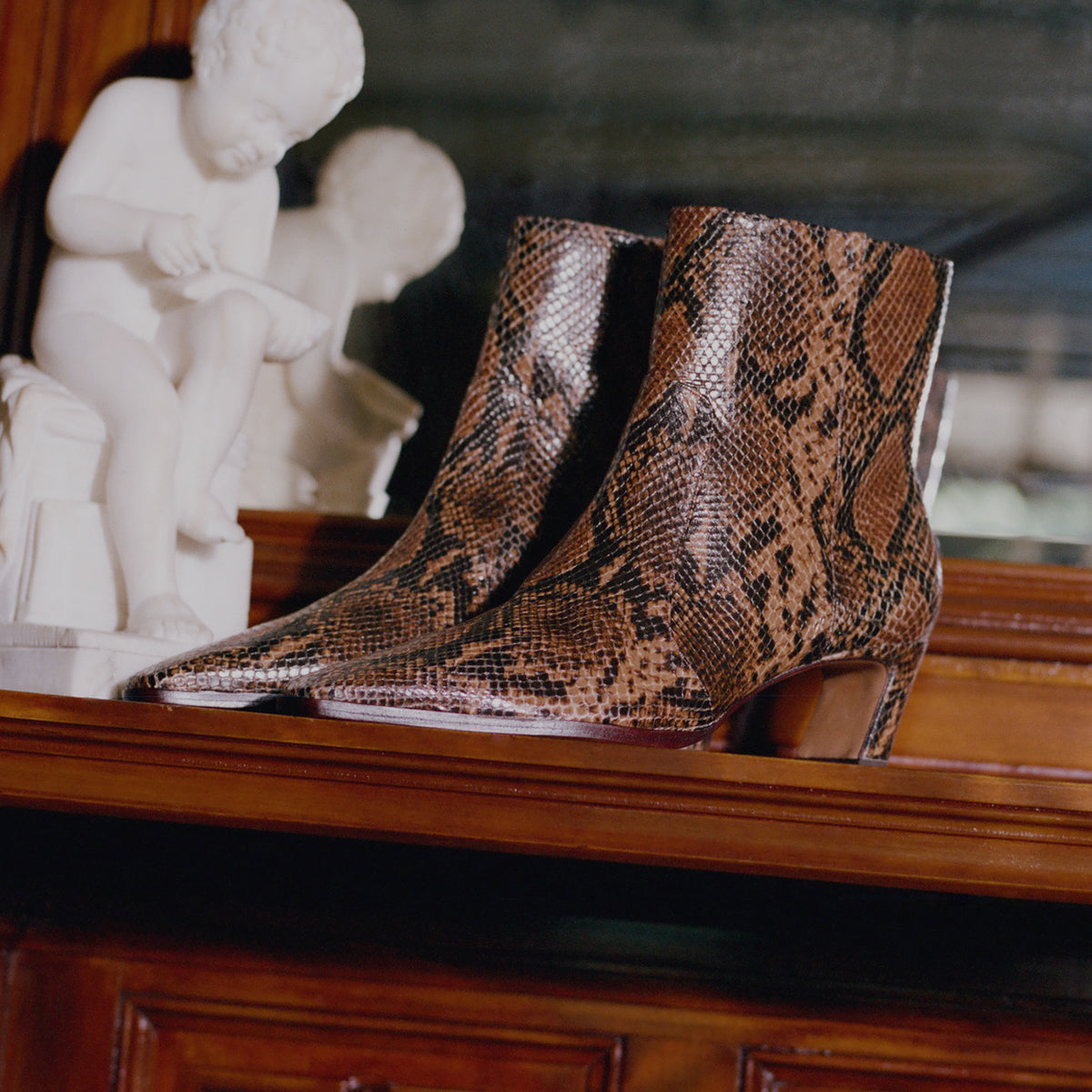 A pair of pointed-toe ankle boots with a brown and black snakeskin pattern, displayed on a wooden mantelpiece next to white classical cherub statues.