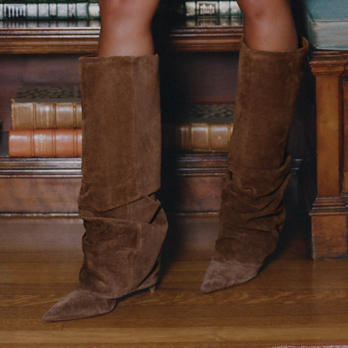 A pair of knee-high brown suede slouch boots with pointed toes, worn by a model standing on a wooden floor in front of a bookshelf.