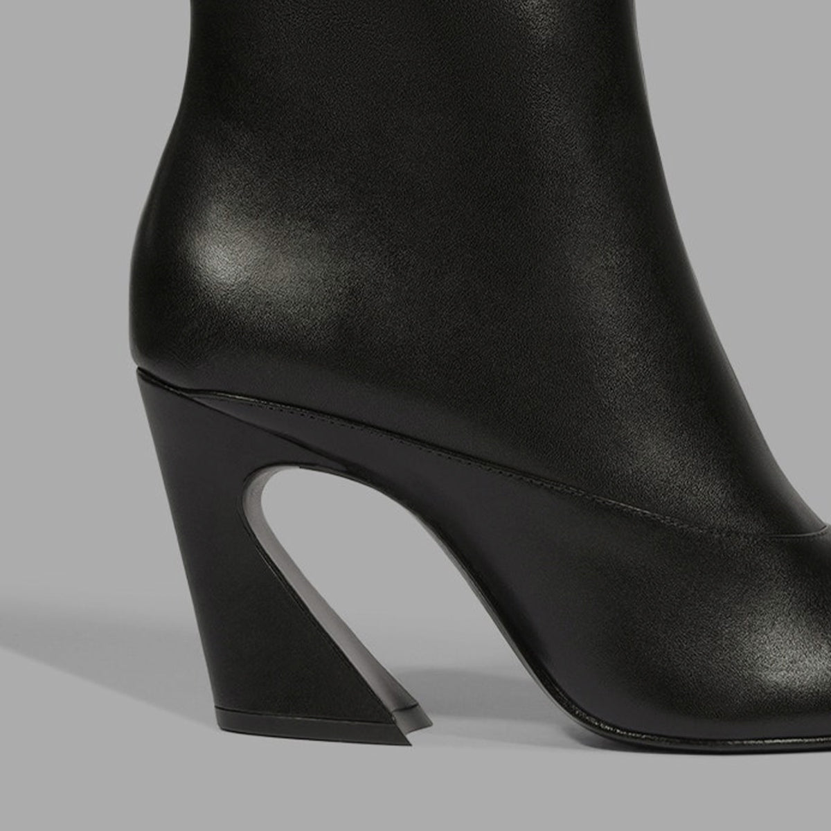 A close-up studio shot of a black leather ankle boot featuring a distinctive sculptural curved block heel on a neutral gray background.