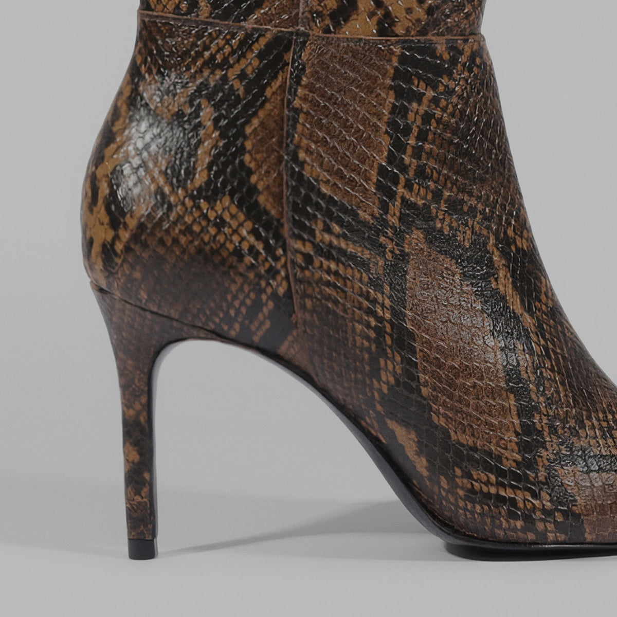 A close-up side view of a high-heeled ankle boot featuring a brown and black snakeskin pattern, set against a neutral gray background.