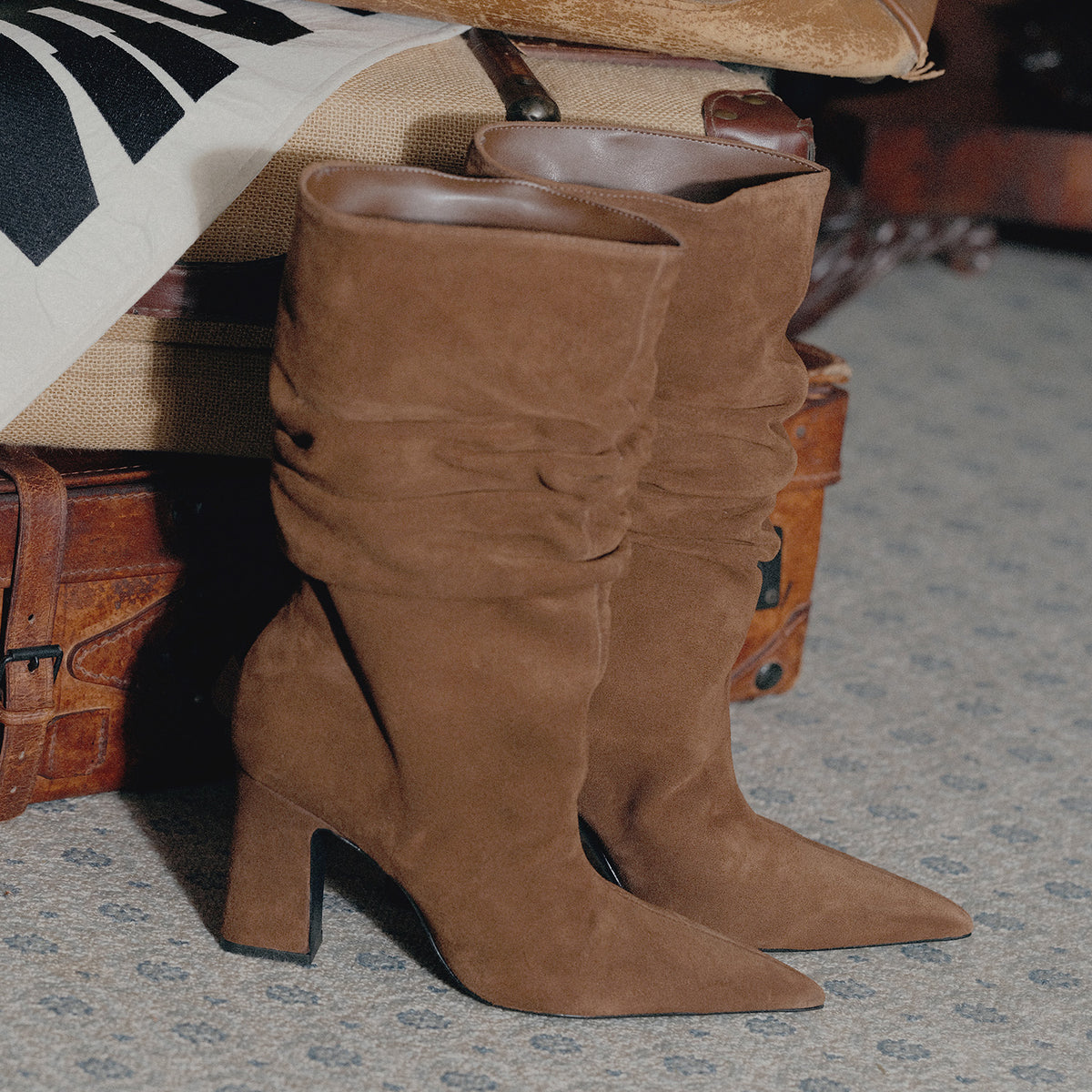 A pair of mid-calf brown suede boots with pointed toes and block heels, styled on a patterned carpet next to vintage leather luggage.