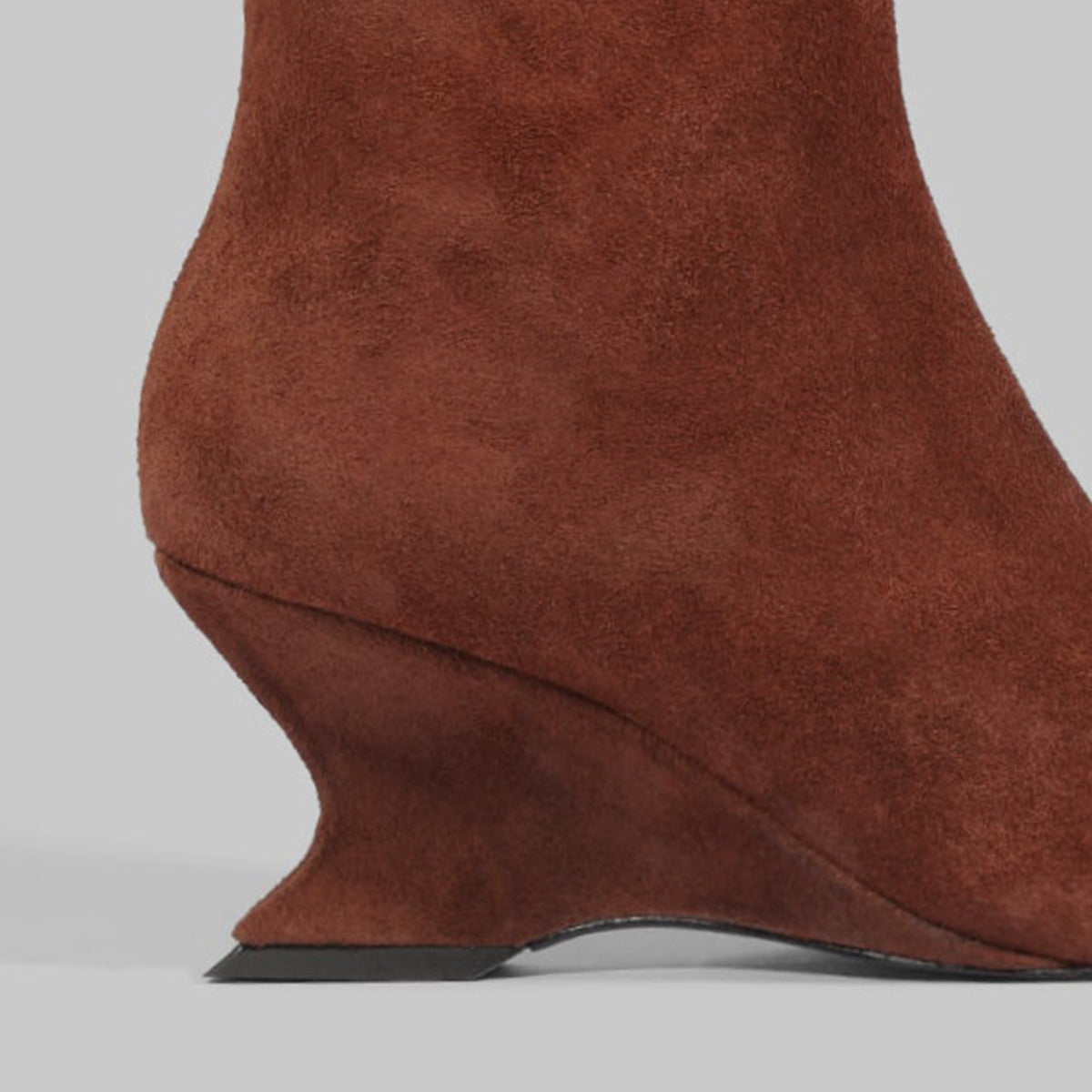 Close-up of a brown suede ankle boot with a sculpted wedge heel, shown against a light gray background.