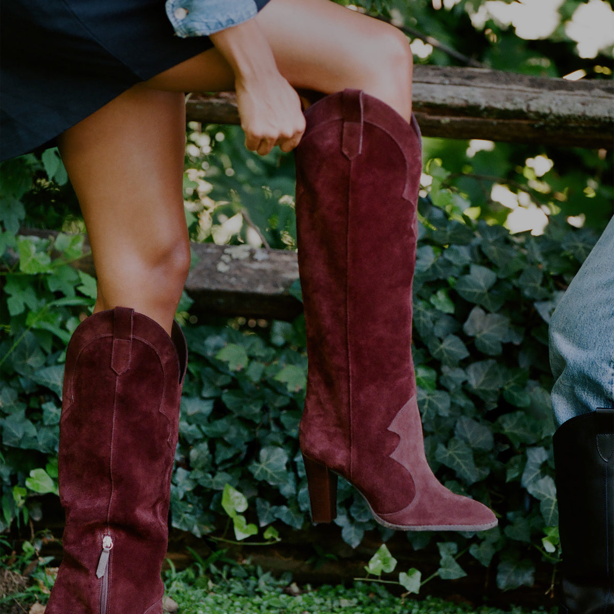 A person wearing tall burgundy suede cowboy boots with mid-calf height heels, sitting on a wooden fence outdoors with green ivy in the background.