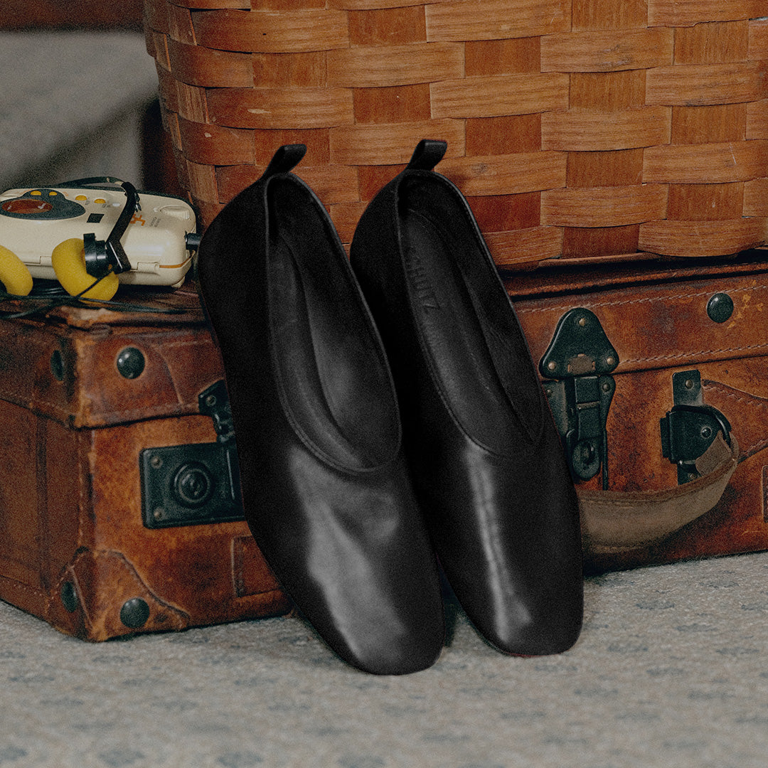 A pair of black leather slip-on shoes with a square toe, styled on top of vintage brown leather suitcases in front of a woven basket.