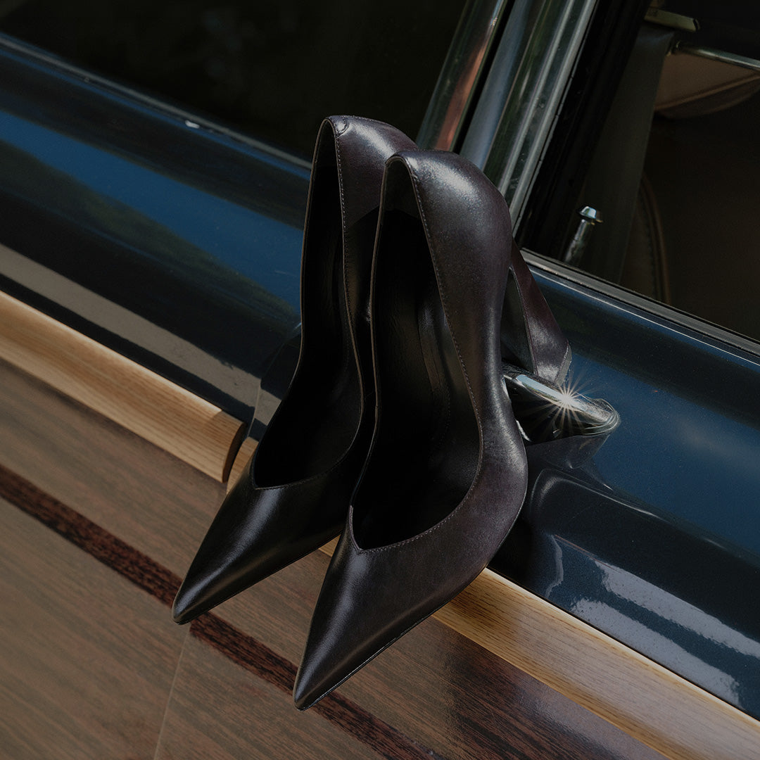 A pointed-toe mule pump with an asymmetrical upper in black leather, placed in front of a vintage suitcase inside a car.