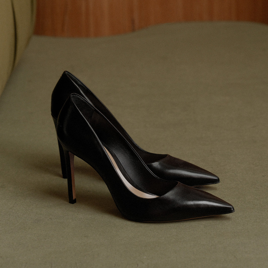 A pair of black leather pointed-toe high heels with slim stiletto heels, displayed on a textured beige surface.