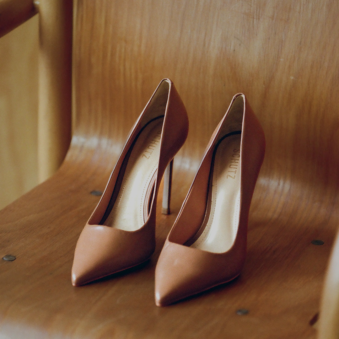 A pair of pointed-toe high heels in a warm tan-brown leather, resting on a wooden chair.